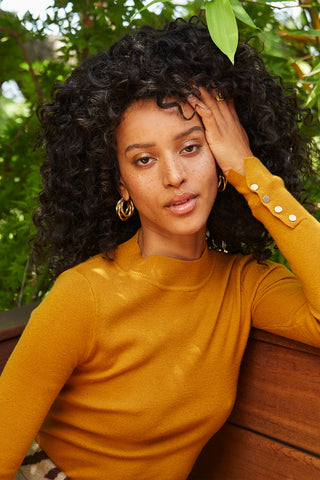 A woman wearing a mustard knit top with sleeve gold snap button detail.